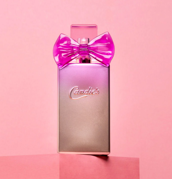 A stylish bottle of Candie's Charm Fragrance, adorned with a glossy pink bow, is set against a pink background. The metallic bottle features the embossed brand name "Candie's" on the front and is elegantly placed on a pink platform.